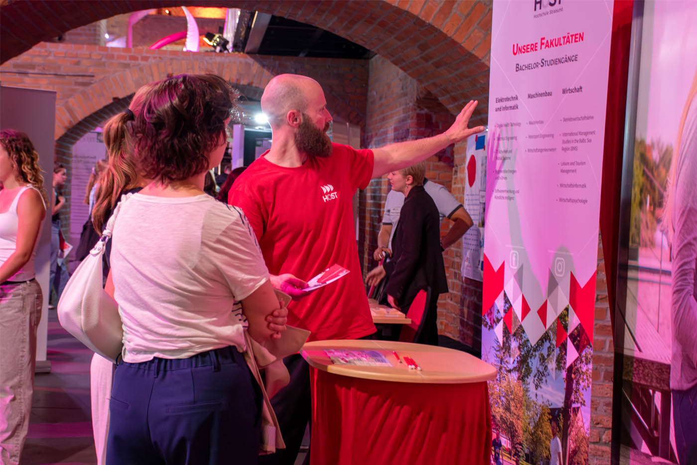 Mann im roten T-Shirt zeigt am Messestand einer Frau etwas auf einem Aussteller-Roll-Up